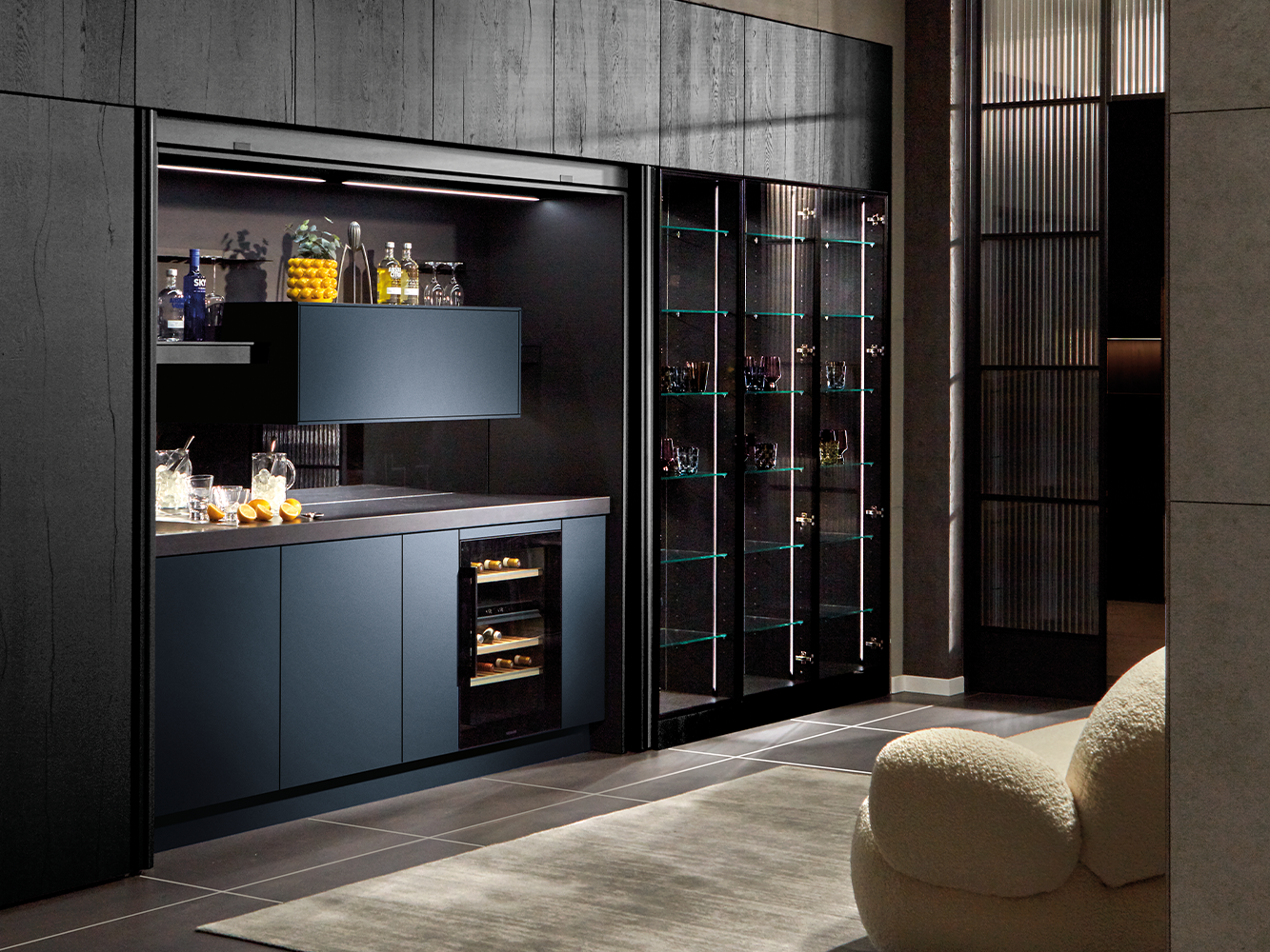 Du siehst eine moderne pronorm Designküche mit integrierter Bar, dunklen Fronten und eleganter Glasvitrine im offenen Wohnraum.