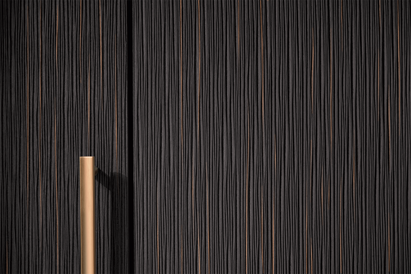 Gros plan sur une façade de cuisine pronorm avec veinage vertical en bois foncé et poignée métallique dorée.