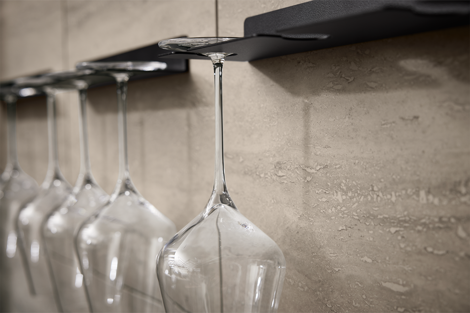 Close-up of wine glasses in black rack mounted on travertine backsplash, featured in walnut and stone handleless kitchen by pronorm