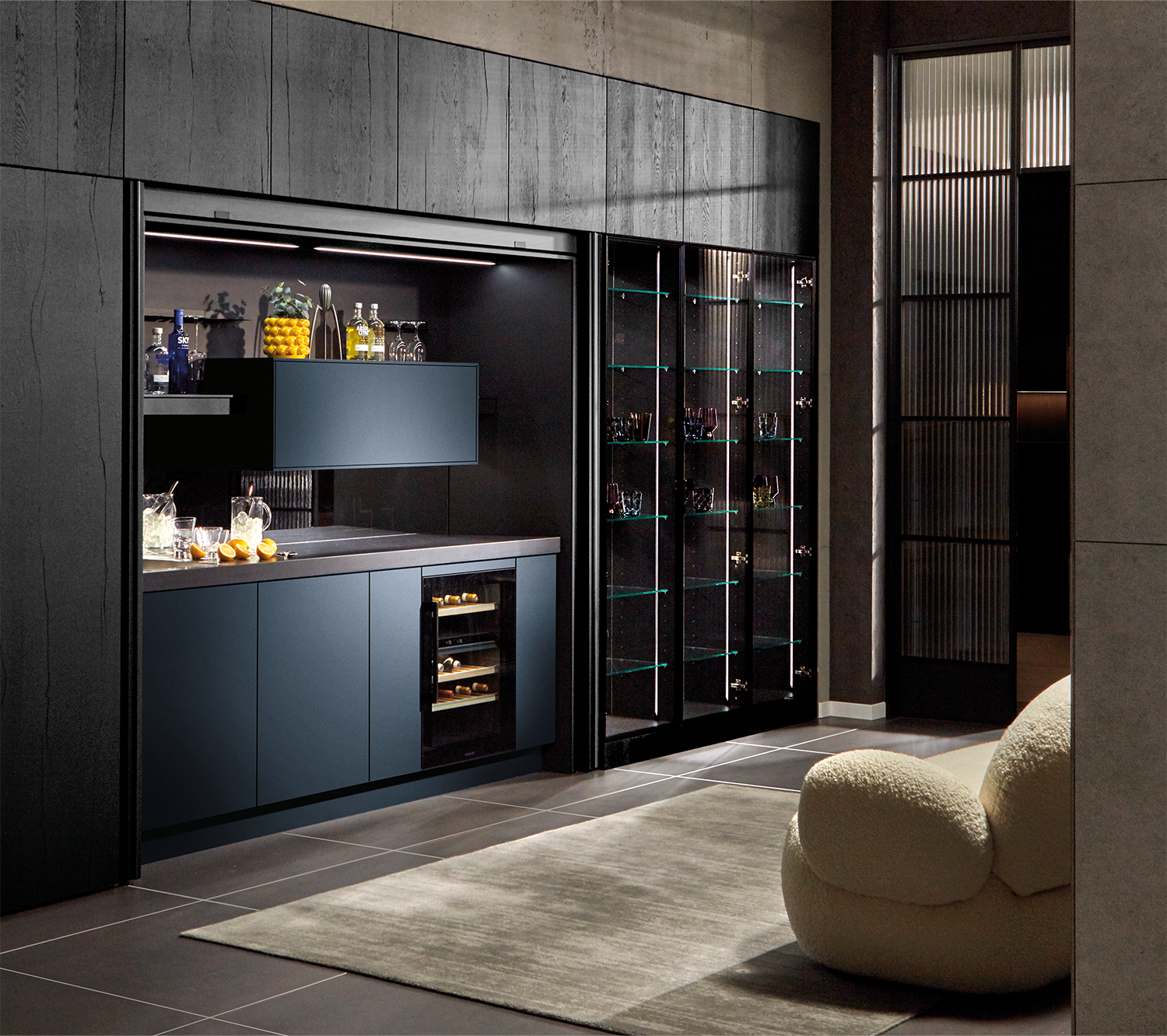 Je ziet een moderne pronorm designkeuken met geïntegreerde bar, donkere fronten en een elegante glazen vitrinekast in een open woonruimte.