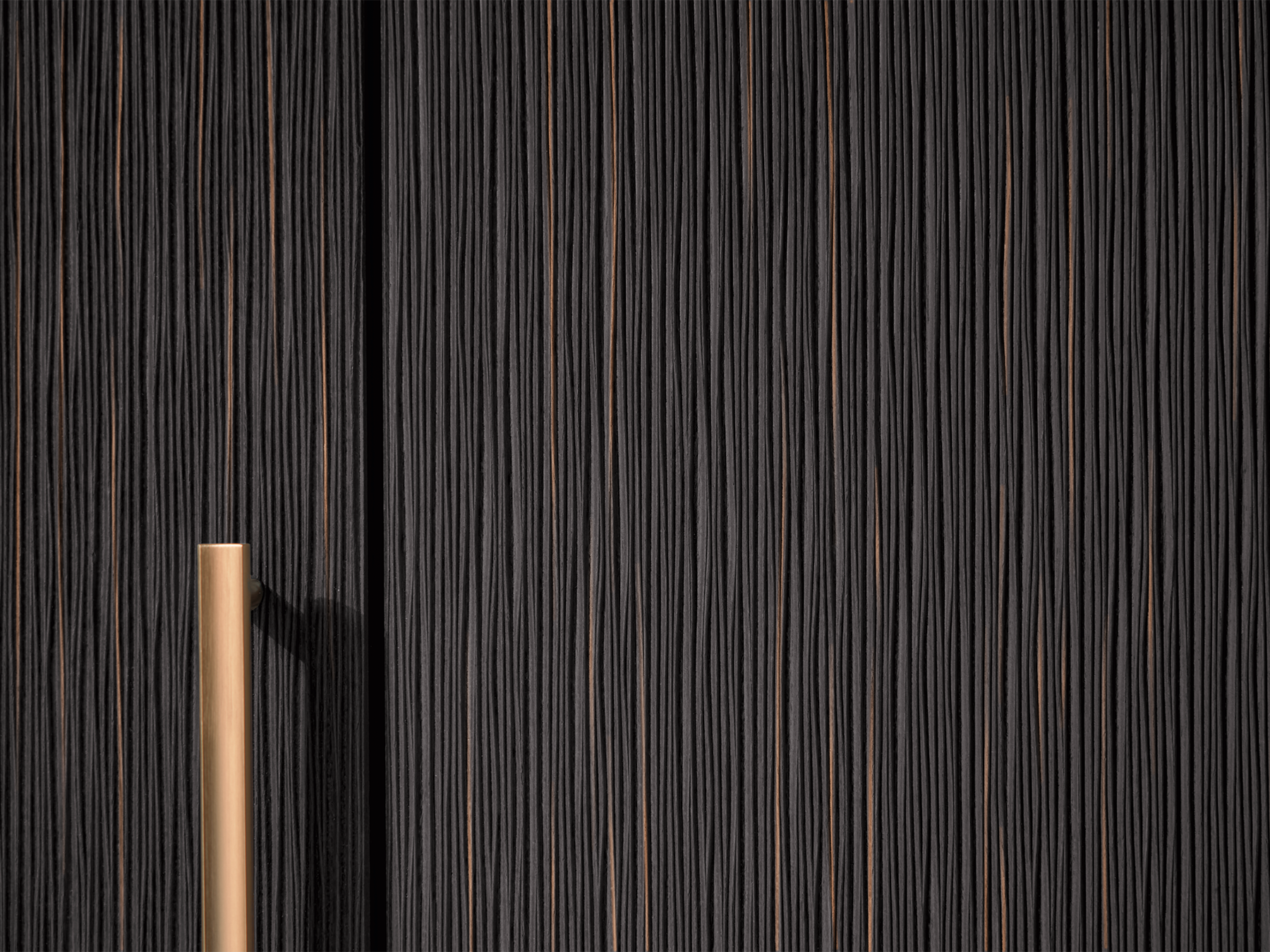 Detailed view of a pronorm kitchen front in amber with a vertically structured surface in a dark wood look and a gold-coloured handle rail for exclusive kitchen design with character.