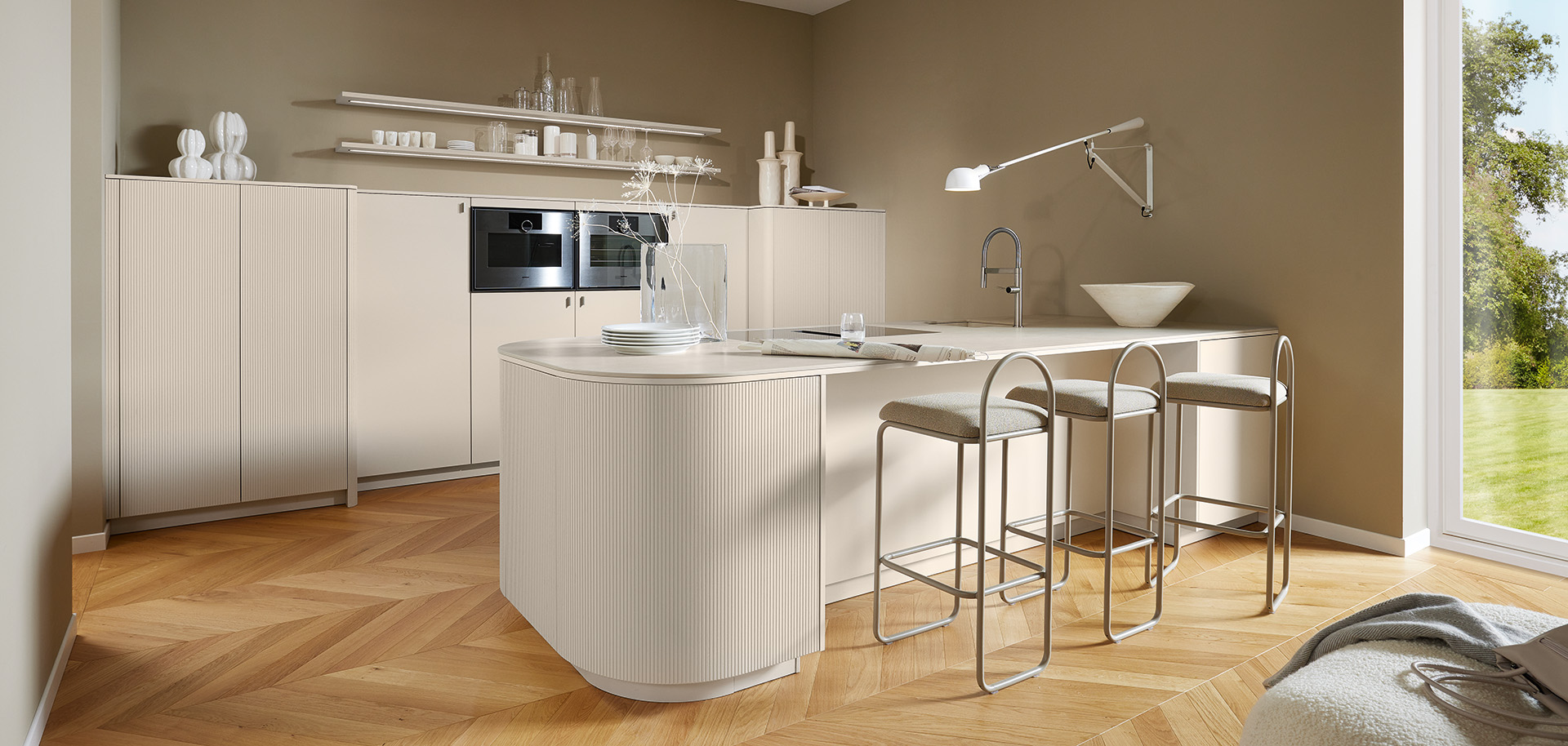 Helle Designküche mit Rillenfront und gerundeter Insel von pronorm in warmen Beigetönen und natürlichem Holzfußboden.