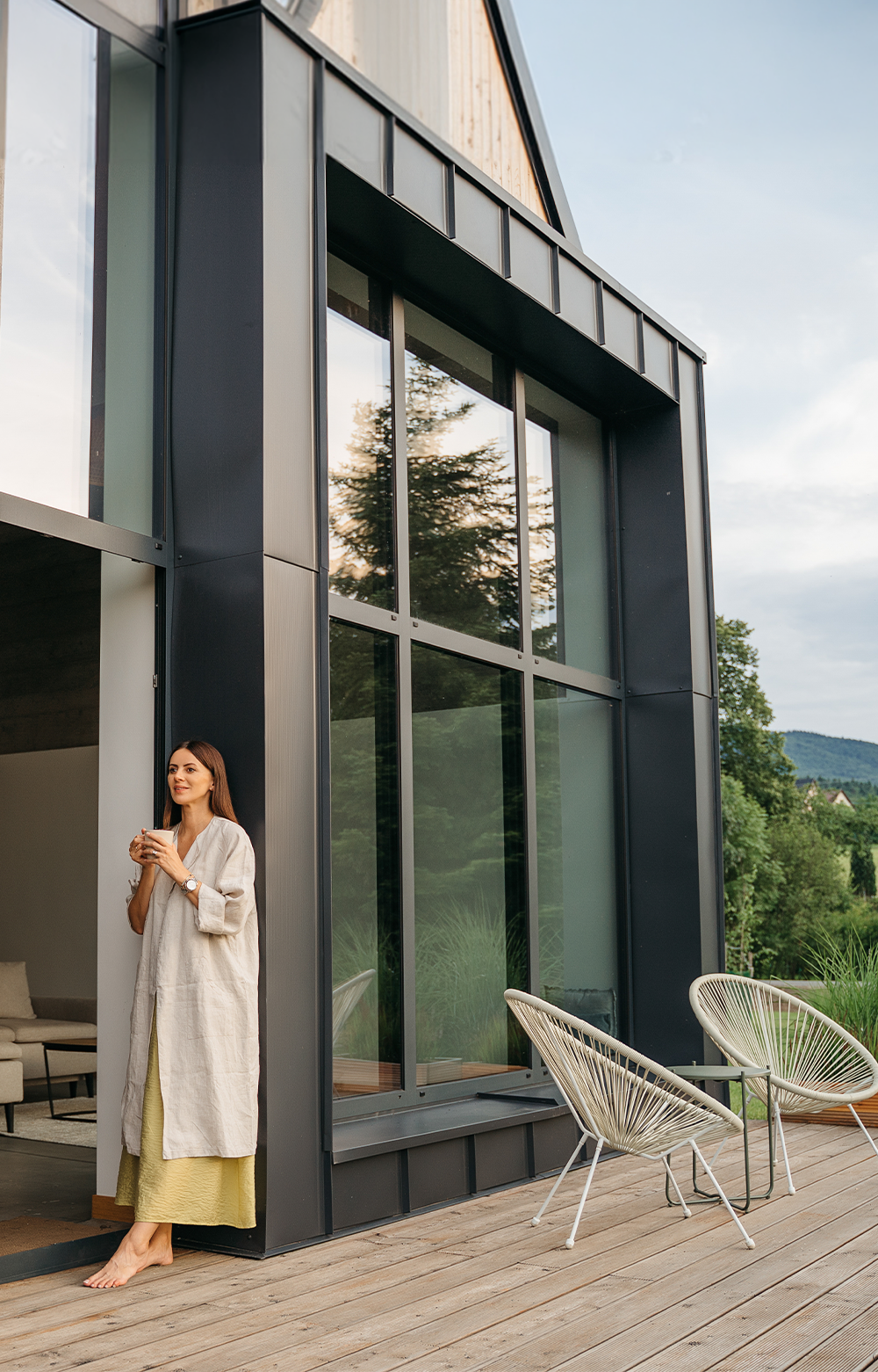 A woman standing on a terrace in front of modern architecture with natural surroundings in the background.